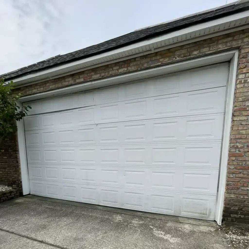 Damaged white panel garage door on a Kenner brick home before replacement